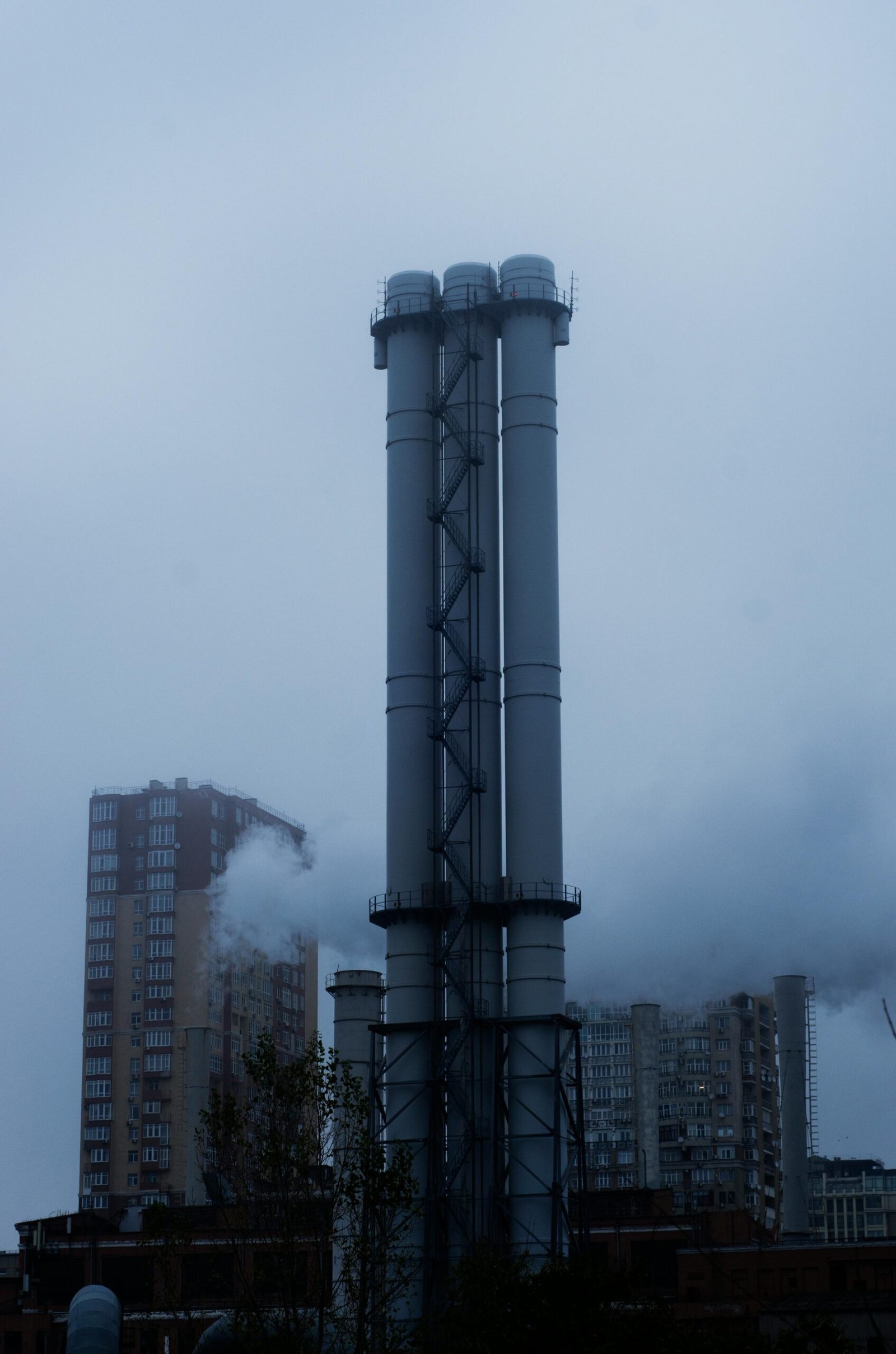 Tall industrial smokestacks emitting smoke against a cloudy urban skyline, highlighting air pollution.
